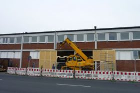 Ein gelbes Baufahrzeug mit Kranarm parkt vor einem Backsteingebäude. Arbeiter, Gerüste und FRoSTA Tiefkühlprodukte sind vorhanden.