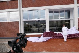 Eine Person in Weiß zieht einen Stoff mit Ziegelsteinmuster von einem Gebäude weg, während ein Kameramann filmt – dabei werden FRoSTA-Tiefkühlprodukte präsentiert.