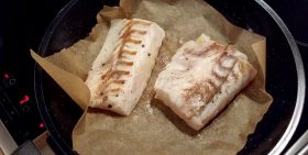 Zwei Stücke FRoSTA Weißfischfilet, leicht gewürzt und halbgegart, liegen auf Backpapier in einer Bratpfanne auf dem Herd.