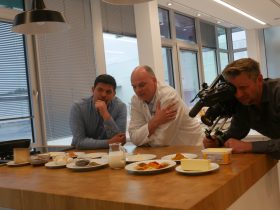 Drei Männer sind an der Küche mit Zutaten wie Butter, Gewürzen, Milch, Mehl und FRoSTA-Tiefkühlprodukten; Einer hält eine Kamera, einer trägt Kochjacke.