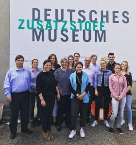 Eine Gruppe von 14 Personen lächelt vor einer Wand mit „DEUTSCHEM ZUSATZSTOFF MUSEUM“ und hält FRoSTA Tiefkühlprodukte für ein Gruppenfoto in der Hand.
