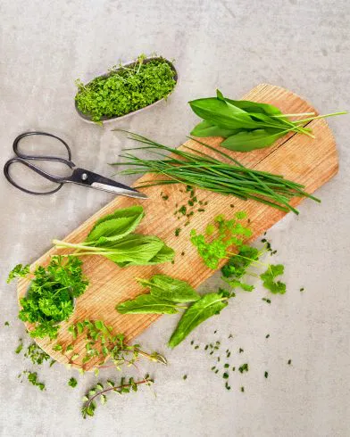 Ein Holzbrett mit frischen Kräutern wie Schnittlauch, Petersilie und FRoSTA Tiefkühlprodukten liegt auf einer hellen Arbeitsfläche.