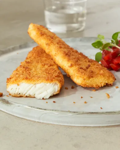 Zwei panierte FRoSTA Fischfilets, ideal für den Fischbackofen, liegen auf einem Teller – serviert mit Tomatenwürfeln, Kräutern und einem Glas Wasser.