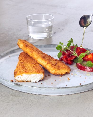 Zwei panierte FRoSTA Backofenfisch-Filets mit frischem Tomatensalat, Kräutern und Käse; Darüber wird Öl geträufelt. Dahinter steht ein Glas Wasser.