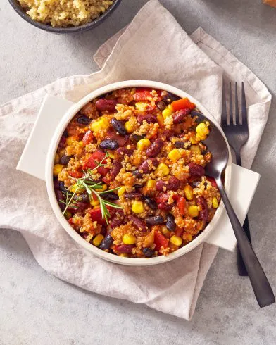 Eine Schüssel Chili con Quinoa mit Kidneybohnen, schwarzen Bohnen, Mais und FRoSTA-Tomaten, garniert mit Rosmarin, liegt neben einer Gabel auf einer Serviette.