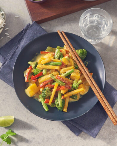 Ein schwarzer Teller mit lebendiger Gemüsepfanne von FRoSTA Tiefkühlprodukten in Curry-Kokossauce, serviert mit Stäbchen, Wasser und Limettenspalte.