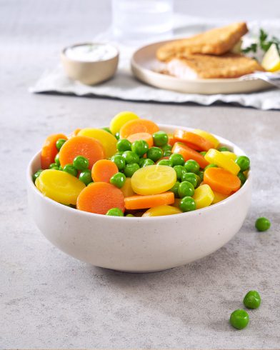 Auf einem hellen Untergrund steht eine weiße Schüssel mit FRoSTA Erbsen und Karotten, im Hintergrund panierte Speisen.