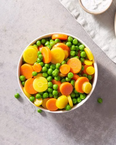 Eine Schüssel mit FRoSTA Tiefkühlprodukten: gelbe und orange Erbsen, Karotten und grüne Erbsen, dazu ein cremiger Dip am Rand.