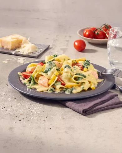 Auf einem Teller liegt cremige Fettuccine mit Spinat, Tomaten und Lachs von FRoSTA. Im Hintergrund Parmesan, Tomaten und ein Glas Wasser.