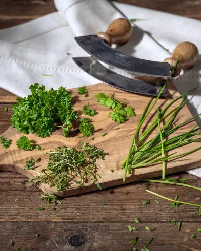 Auf dem Tisch steht ein Holzbrett mit frischen Gartenkräutern und FRoSTA-Tiefkühlprodukten. Dahinter ein Kräutermesser und ein weißes Tuch.