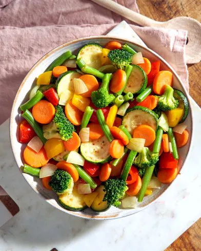 Eine Schale FRoSTA italienisches Tiefkühlgemüse – Karotten, Brokkoli, Zucchini, Bohnen, Zwiebeln und Paprika – auf weißem Marmor.
