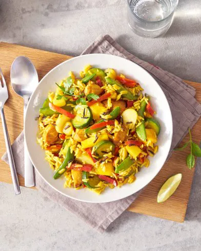 Auf einem weißen Teller liegt buntes FRoSTA-Gemüsereis mit Kräutern, dazu Limette, Minze und Wasser auf hellem Tisch. Curryduft liegt in der Luft.