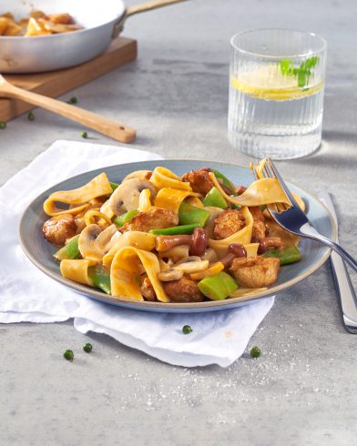 Ein Teller mit FRoSTA Tiefkühlpasta, Hähnchen, Pilzen, Paprika und Sauce steht neben Besteck und einem Glas Wasser mit Zitrone.