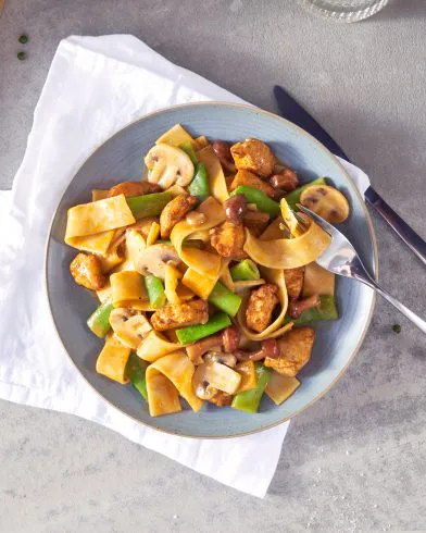 Ein Teller breite Nudelnudeln mit geschnetzeltem Hähnchen, Pilzen, grünen Paprika und Bohnen in heller Soße, zubereitet mit FRoSTA-Tiefkühlprodukten.