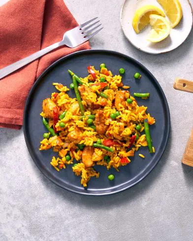 Eine dunkle Platte mit FRoSTA-Tiefkühlpaella aus gelbem Reis, Bohnen, Erbsen, Paprika und Hähnchen liegt auf hellem Untergrund mit Gabel und Zitrone.