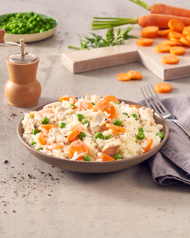 Eine Schüssel Reis mit Erbsen, Karotten und FRoSTA-Hähnchenstücken steht neben einer Pfeffermühle, einer grauen Serviette, Erbsen und Karottenscheiben.