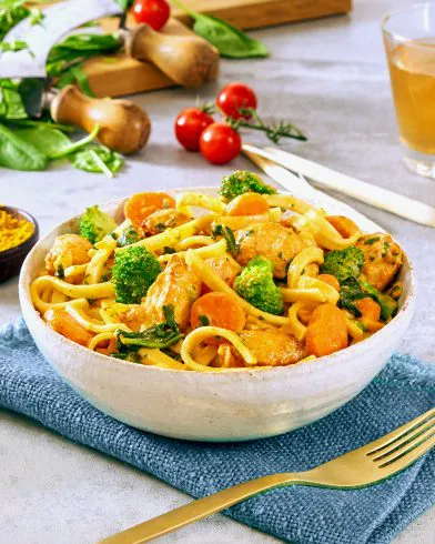 Eine Schüssel mit FRoSTA Tiefkühlprodukten mit Nudeln, Hähnchen, Brokkoli, Möhren und Currysoße steht auf blauem Tuch mit Beilagen im Hintergrund.