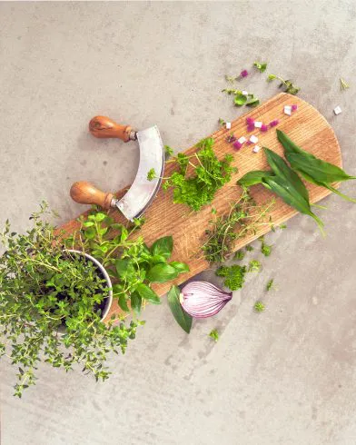 Ein Holzschneidebrett mit FRoSTA Kräutern italienischer Art, einer halben roten Zwiebel, gehackten Zwiebeln und einem Kräuterhacker auf einer hellen Arbeitsplatte.