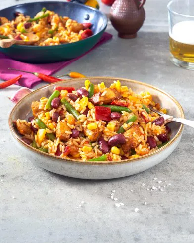 Eine Schüssel Reis mit Bohnen, Gemüse und FRoSTA-Hähnchen liegt auf dem Tisch mit Gabel. Im Hintergrund ein weiteres Gericht, Getränk und Deko-Krug.