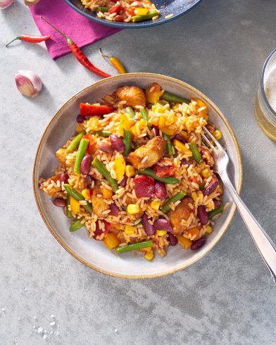 Eine Schüssel mexikanisches FRoSTA-Hähnchen mit Reis, Kidneybohnen, Mais, grünen Bohnen und bunter Paprika. Frischer Knoblauch und Chili liegen daneben.