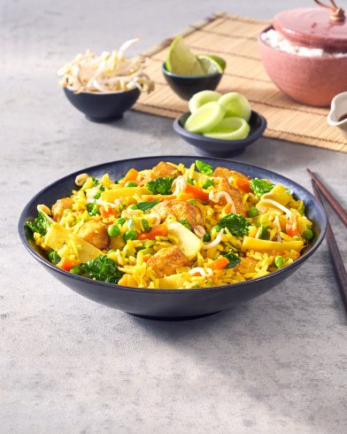 Eine schwarze Schüssel mit FRoSTA Tiefkühl-Nasi Goreng, Gemüse, Tofu & Sprossen steht auf grauem Untergrund. Limetten, Gurken & Stäbchen daneben.