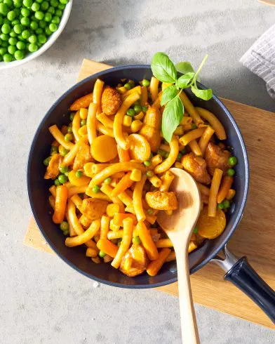 Eine Pfanne mit leckeren FRoSTA Tiefkühlprodukten: Nudelpfanne mit Hähnchen, Erbsen, Karotten, gelb-orange Soße und frischem Basilikum.