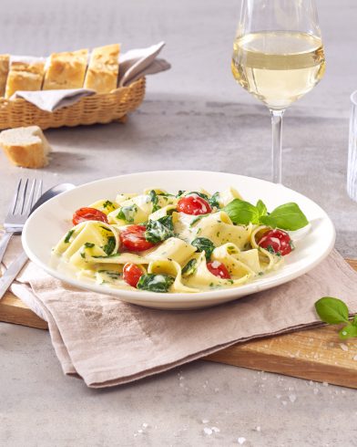 Ein Teller FRoSTA Pappardelle Crème Spinaci mit cremiger Sauce, Spinat und Tomaten wird neben Brot und Wein serviert.