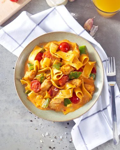 Eine Schale FRoSTA Tiefkühlpasta mit Hähnchen, Paprika, Cherrytomaten und cremiger Soße, dazu ein Glas Orangensaft und Besteck.