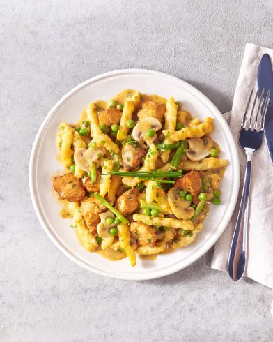 Ein weißer Teller mit FRoSTA-Nudeln in Sahnesauce, Champignons, Erbsen, gebratenem Hähnchen und Schnittlauch. Gabel und Messer auf einer Serviette daneben.
