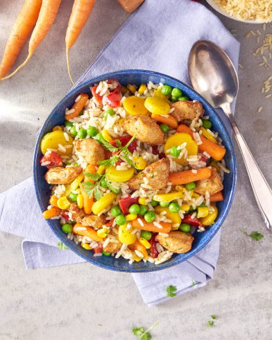 Eine blaue Schüssel mit FRoSTA Reispfanne mit Hähnchen – Reis, Hühnchen, Erbsen, Mais, Karotten und rote Paprika – auf einem lavendelfarbenen Tuch mit einem Löffel.