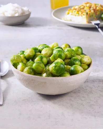 Eine beige Schüssel gefüllt mit gedämpftem FRoSTA-Tiefkühlrosenkohl steht auf hellem Untergrund. Im Hintergrund sind weitere Teller zu sehen.