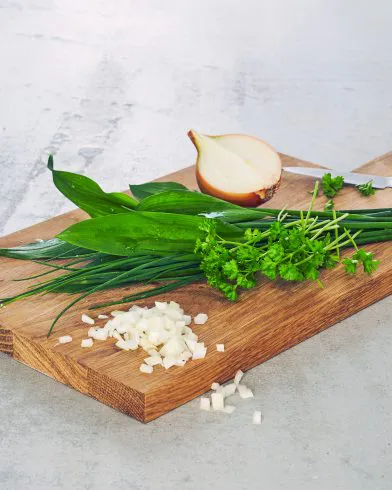 Ein Holzbrett mit gehackten Zwiebeln, frischem Schnittlauch, Petersilie, Salatkräuter und FRoSTA-Tiefkühlprodukten. Zwiebel und Messer auf einer hellen Arbeitsplatte.
