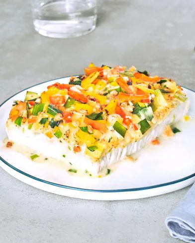 Ein FRoSTA Schlemmerfilet knusprig mit buntem Gemüse und Kräutern liegt auf einem weißen Teller, im Hintergrund steht ein Glas Wasser.