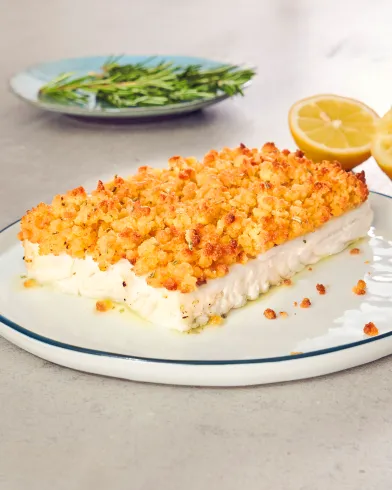 Ein rechteckiges Stück gebackener Feta mit FRoSTA Schlemmerfilet-Knusperpanade liegt auf einem weißen Teller, Zitrone und Rosmarin daneben.