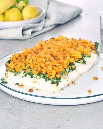 Ein gebackenes FRoSTA Schlemmerfilet knusprig mit goldener Kruste liegt auf weißem Teller, dazu gelbe Kartoffeln im Hintergrund.