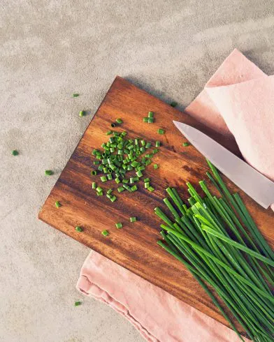 Ein Holzschneidebrett mit frischem Schnittlauch, etwas gehackt und einem Küchenmesser – perfekt für FRoSTA Tiefkühlprodukte. Rosa Stoff darunter.