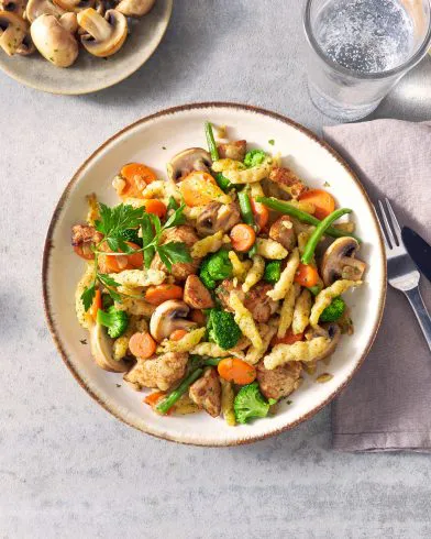 Ein Teller FRoSTA Spätzle-Pfanne mit Pilzen, Karotten, Brokkoli, Bohnen und Fleisch, garniert mit Petersilie. Besteck und Wasserglas daneben.
