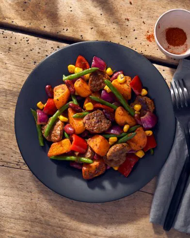 Ein Teller mit sautierten FRoSTA Tiefkühlprodukten: Kartoffeln, grüne Bohnen, Mais, Kidneybohnen, Paprika und Zwiebeln steht neben Paprika und Besteck auf einem Tisch.