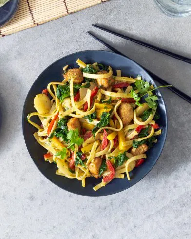 Ein schwarzer Teller mit gebratenen Nudeln, Tofu, Paprika, Gemüse und Koriander – ideal zu FRoSTA würzigen Thai-Hähnchen. Essstäbchen auf hellgrauer Oberfläche.