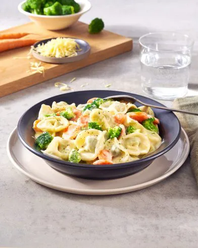 Eine Schale FRoSTA-Tiefkühl-Tortellini in Käse-Sahne-Sauce mit Brokkoli und Karotten, dazu Wasser, Gabel, Käse, frisches Gemüse.