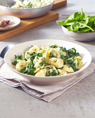 Eine Schüssel FRoSTA Tiefkühl-Tortellini mit Spinat in cremiger Sauce steht auf einer Serviette, daneben frischer Spinat und Käse.