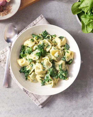 Eine weiße Schüssel gefüllt mit Tortellini mit Spinat von FRoSTA wird auf einer grauen Serviette serviert, daneben liegt ein Löffel auf einer hellgrauen Unterlage.