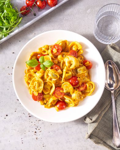 Ein weißer Teller mit FRoSTA-Tortellini mit Kirschtomaten, Basilikum und Sahnesauce, serviert mit Rucola und gerösteten Tomaten als Beilage.