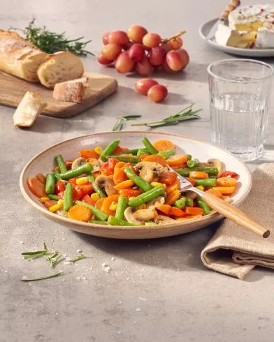 Ein Teller FRoSTA Tiefkühl-Gemüsepfanne Provence mit grünen Bohnen, Karotten und Pilzen steht auf dem Tisch, dazu Brot und Käse.