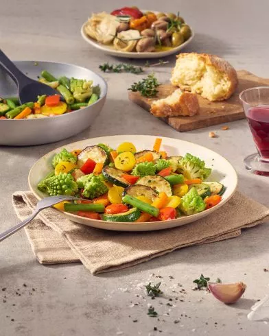 Ein Teller bunter italienischer FRoSTA-Gemüsepfanne steht auf einer Serviette. Daneben Gemüse, Getränke, Brot und Artischocken mit Pickles.