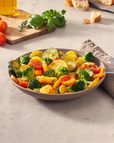 Eine Schüssel bunter, dampfender FRoSTA-Tiefkühlgemüse à la Toscana mit Brot, Kräutern und Serviette steht auf dem Tisch.