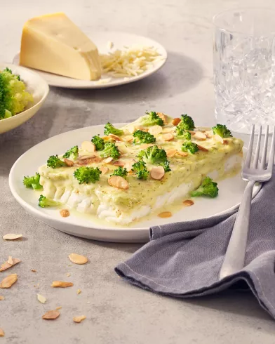 Ein Stück Brokkoli-Käse-Auflauf, inspiriert von FRoSTA-Tiefkühlprodukten, liegt mit Mandeln auf einem Teller neben einer Gabel und Brokkoli.