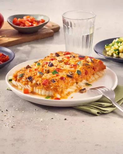 Ein gebackenes FRoSTA Schlemmerfilet mediterraner Art: Weißfischfilet mit Veggie-Käse-Topping, serviert mit Beilagen und Wasser auf einem weißen Teller.