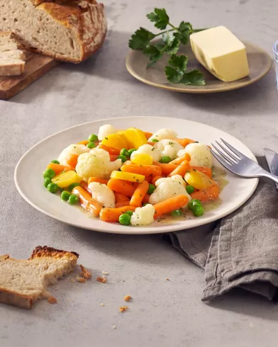 Ein Teller FRoSTA-Tiefkühl-Sommergemüse – Karotten, Erbsen und Blumenkohl – liegt mit Brot, Butter und Petersilie auf dem Tisch.