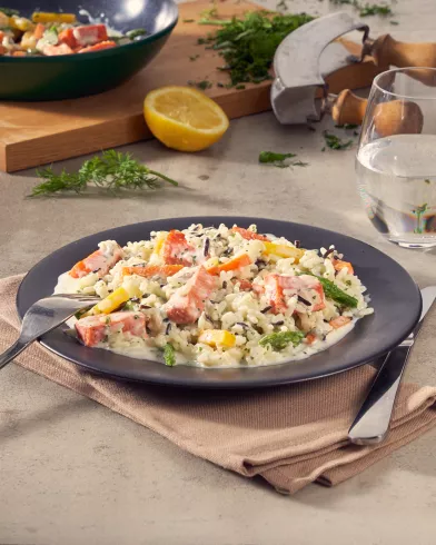 Ein Teller mit cremigem Reis, gewürfeltem FRoSTA Wildlachs, gelber Paprika und Kräutern steht neben Gabel, Messer und Wasserglas; Zitrone und Kräuter im Hintergrund.